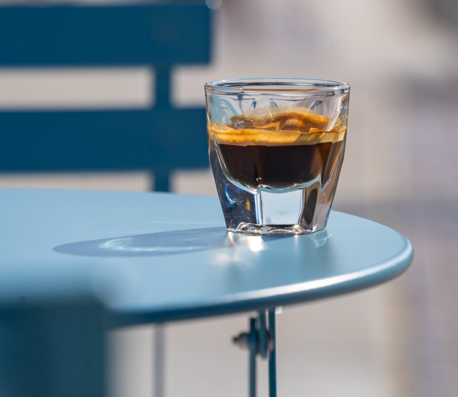 A shot of espresso in a glass on a blue café table in the sun